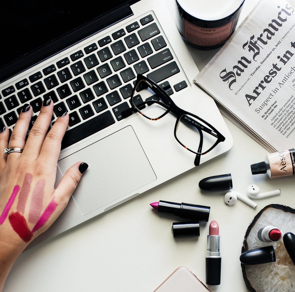 A desk flat lay with an open laptop, lipstick swatches on the back of a hand, tortoiseshell glasses, a newspaper and a candle