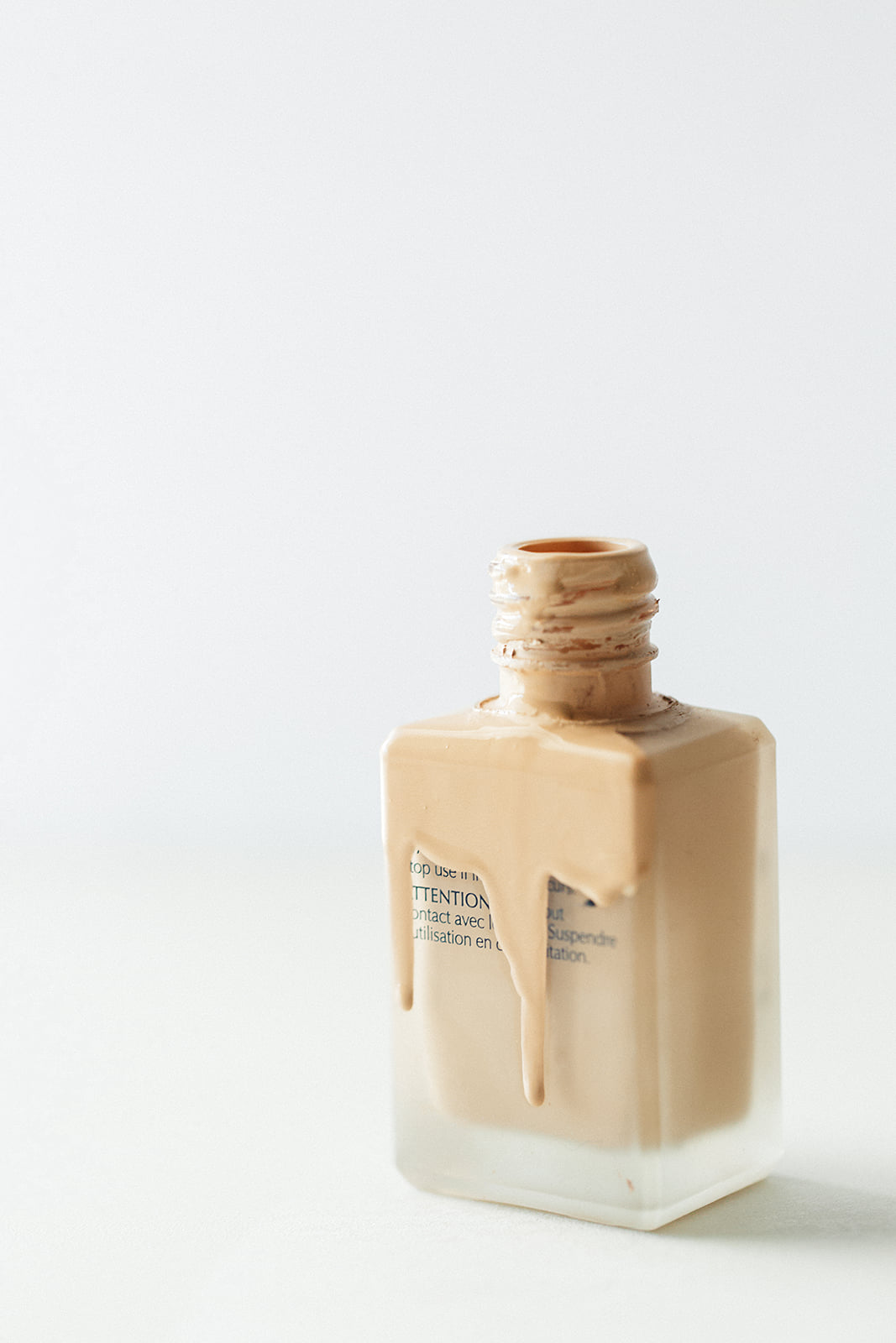 An open frosted glass bottle of liquid foundation with cream dripping down the front on a white background