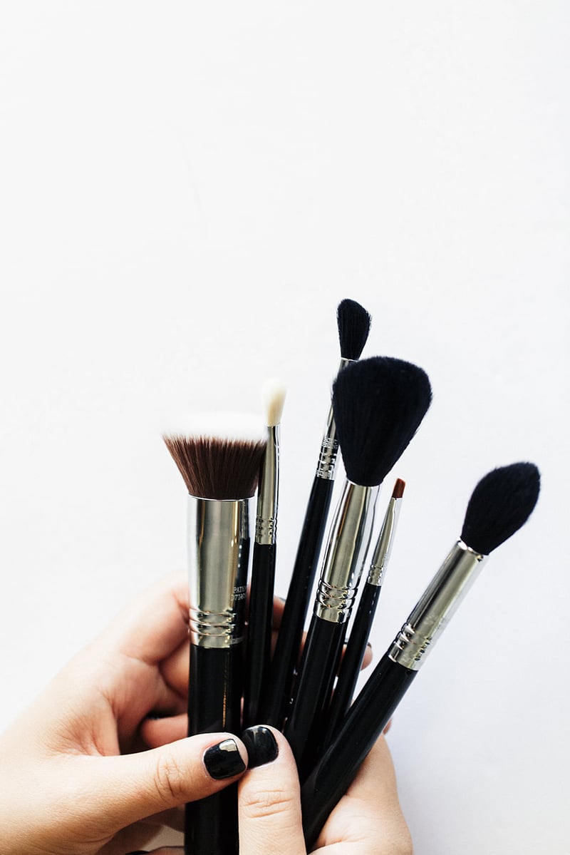 Two hands with glossy black nails holding a bunch of makeup brushes upright against a white background