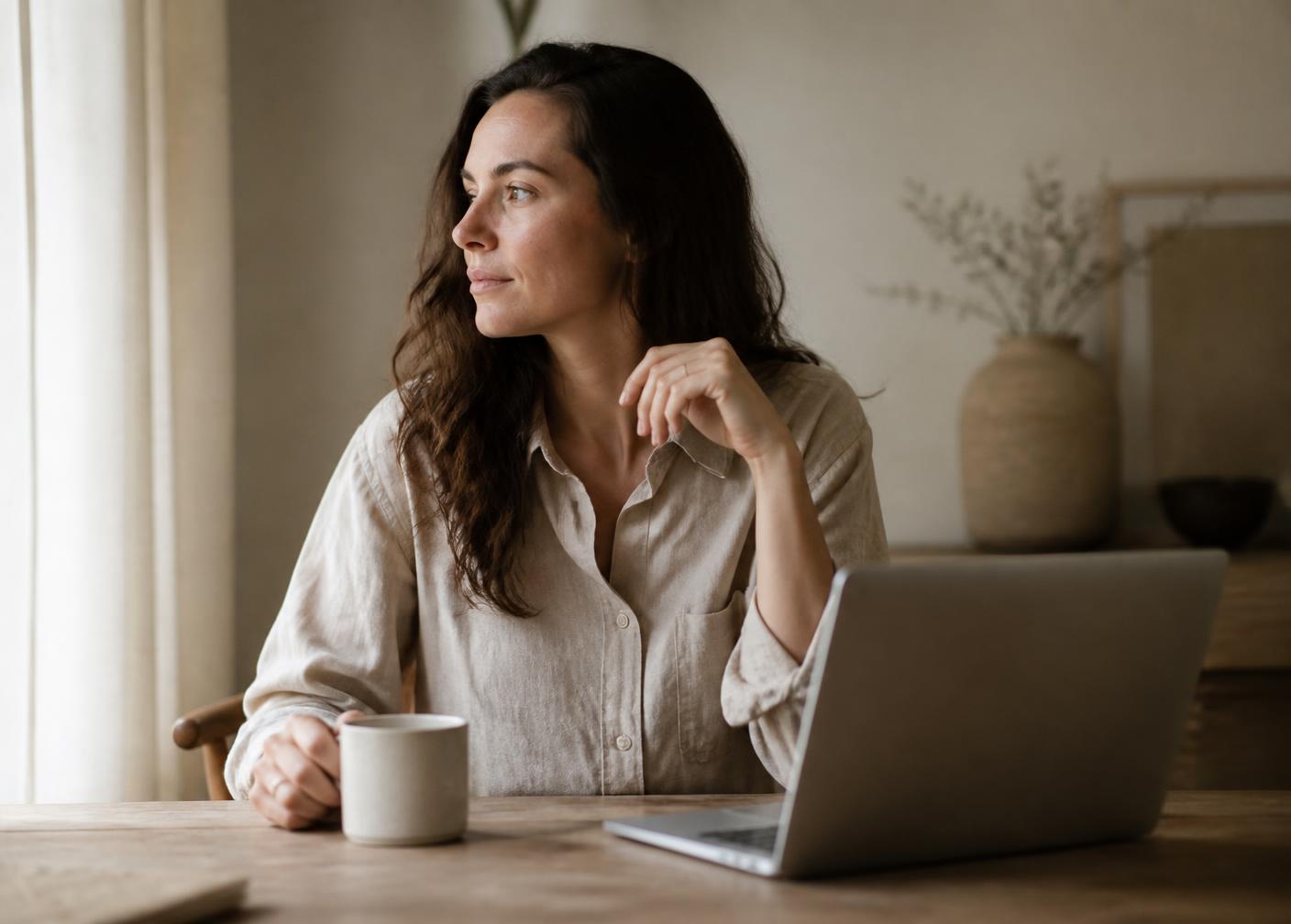 A mother sitting at a sunlit table with a laptop and a cup of coffee